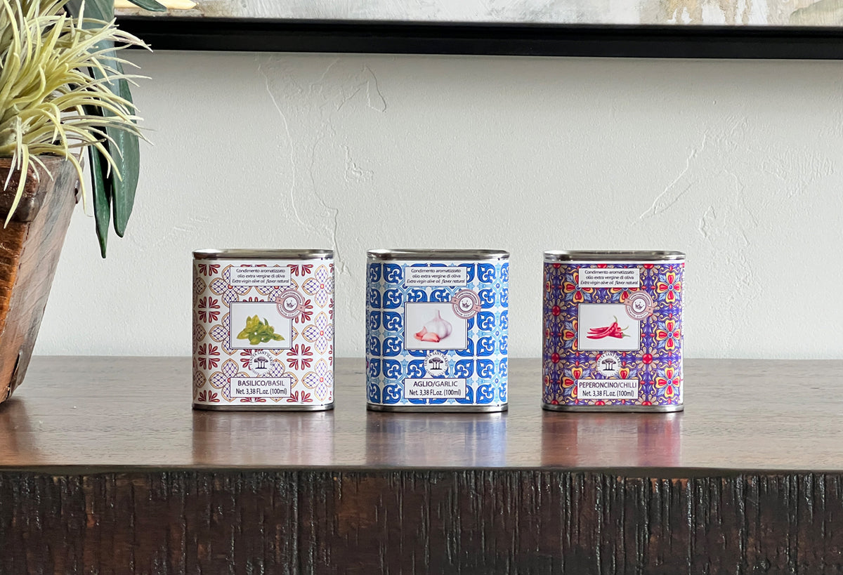 Three decorative metal tins with patterned labels in red, blue, and multicolor designs containing infused olive oil.