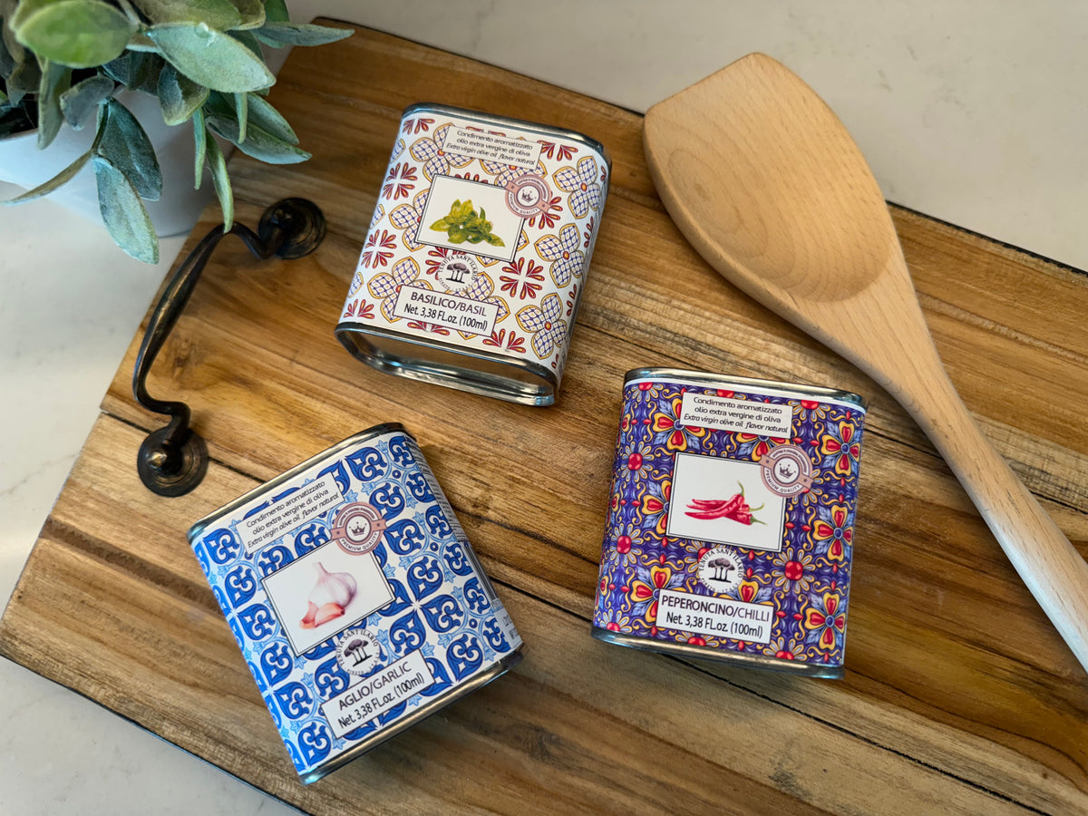 Three decorative olive oil tins and wooden spoon arranged on a wooden board with fresh herbs and black utensil.