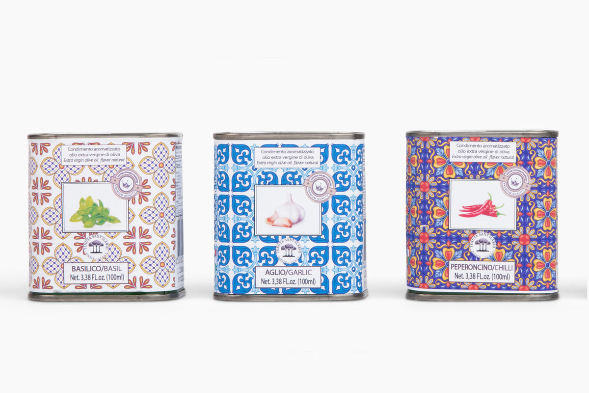 Three cylindrical tins with decorative patterned labels containing basil, garlic, and pepper infused olive oils.
