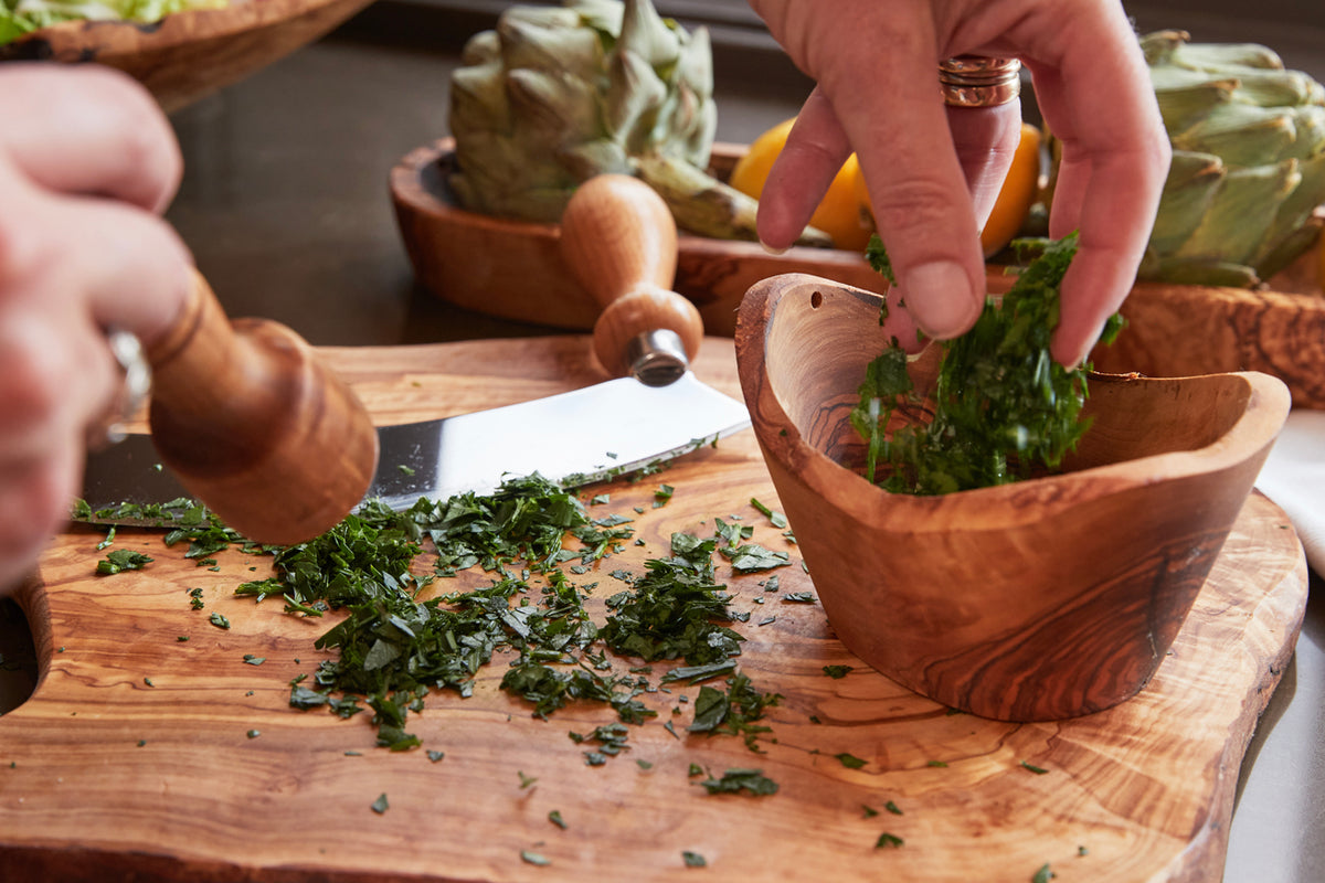 Wooden serving bowl and mortar with pestle used for grinding fresh herbs on a wooden board.