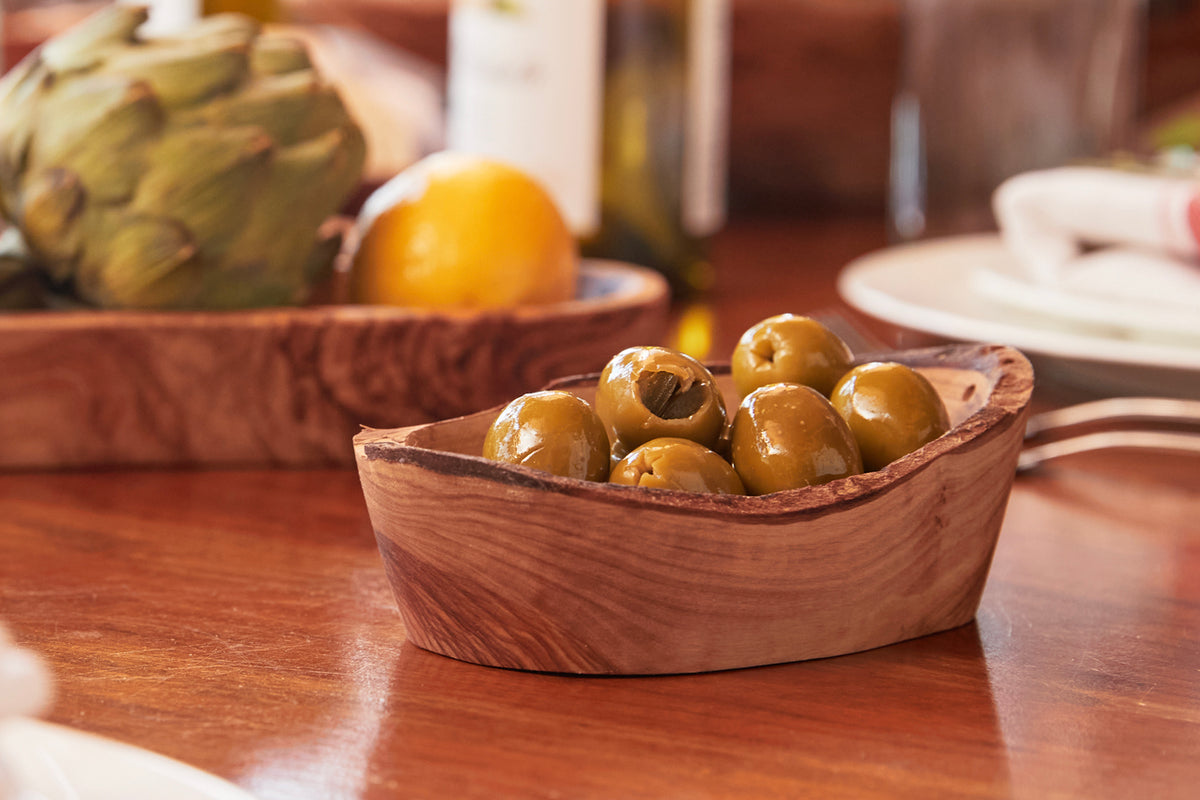 Wooden serving bowl filled with green olives on a dining table with bread and citrus.