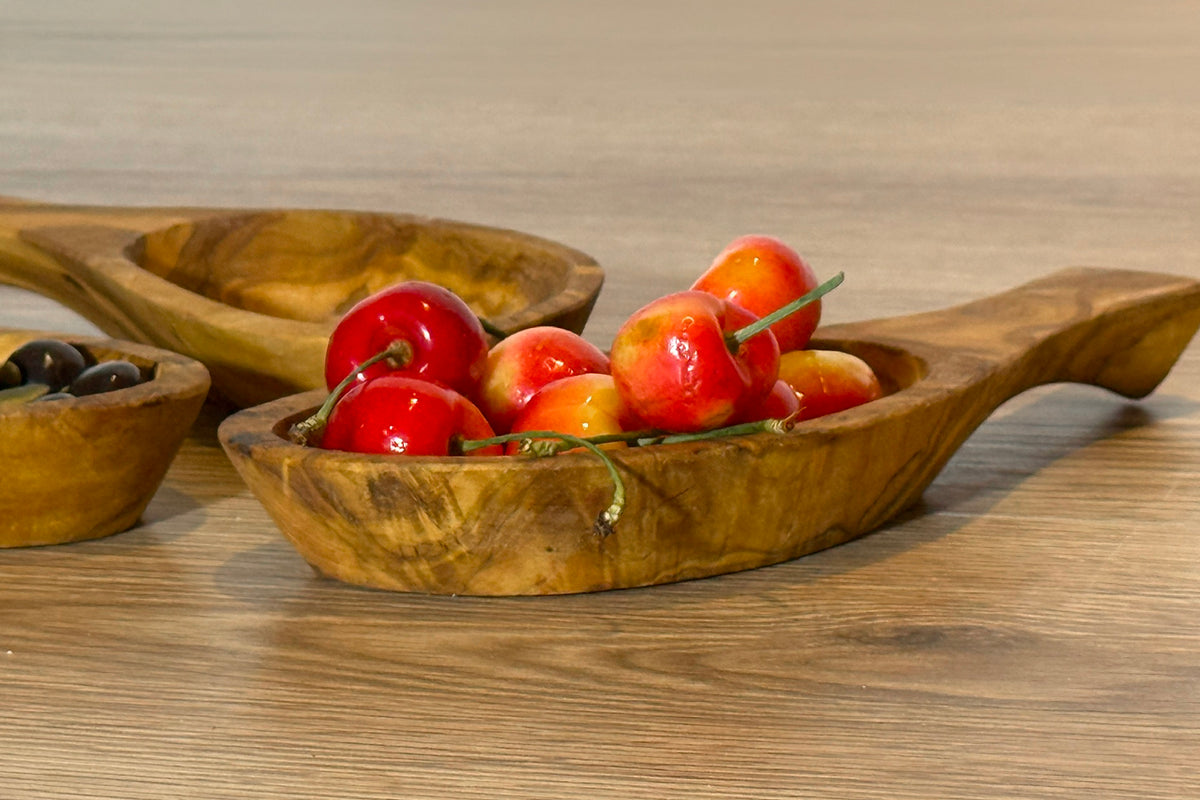 Wooden bowls with handles holding red peppers and black olives on a wooden surface.