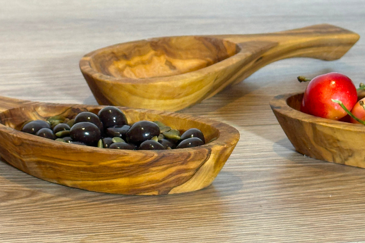 Wooden olivewood bowls with handles containing olives and tomatoes on white surface.