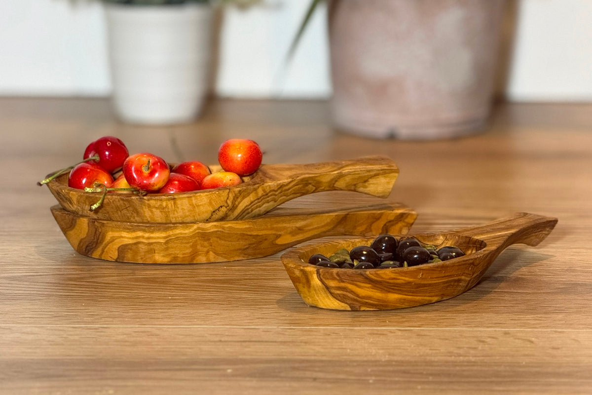 Wooden olive-wood bowls with handles, one containing red tomatoes and another with dark olives.