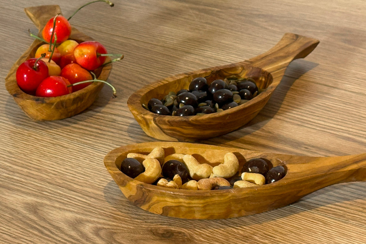 Wooden bowls with handles holding olives, nuts, and cherry tomatoes on a wood surface.