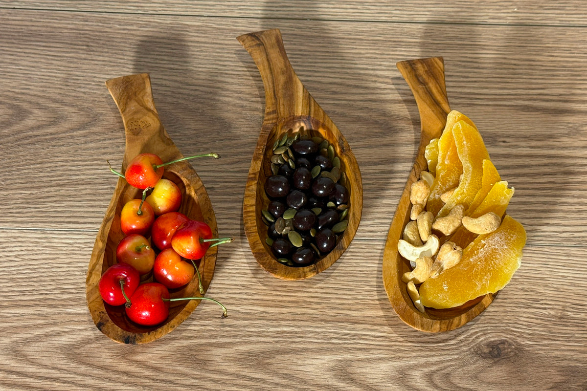 Three wooden bowls with handles holding red tomatoes, black olives, and lemon slices on wood surface.