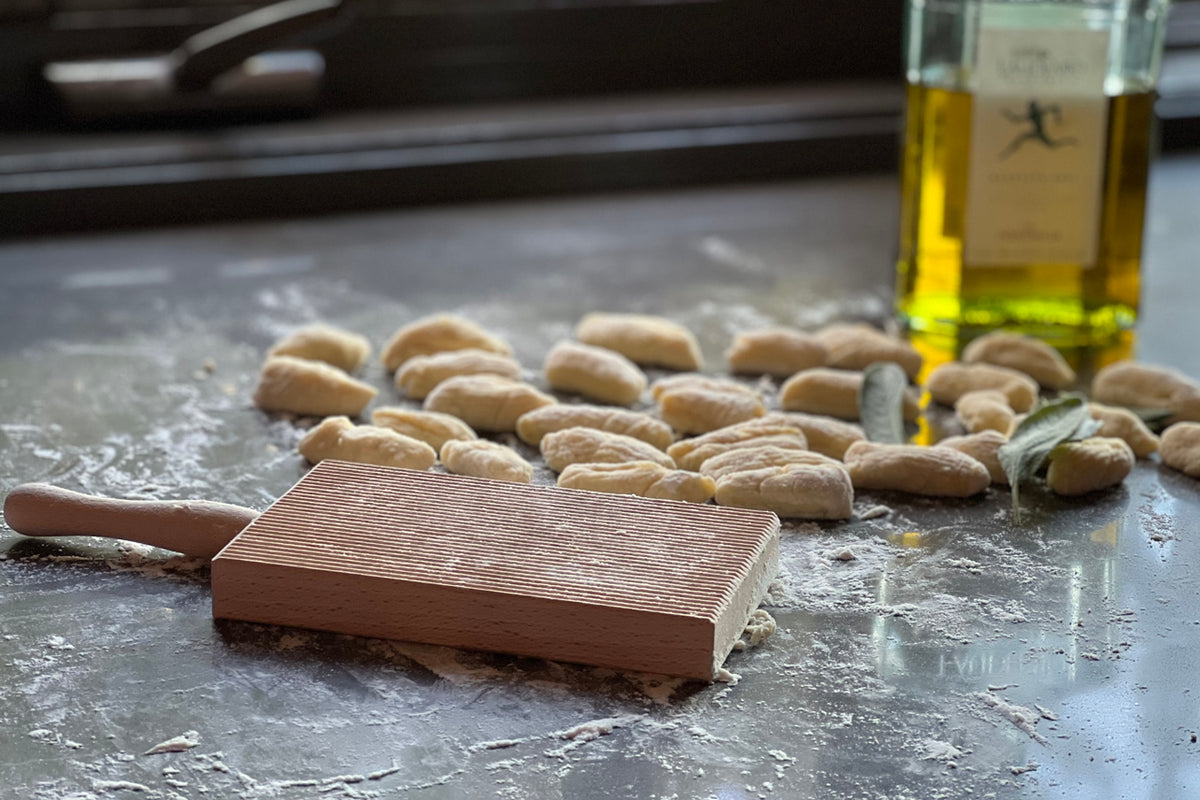 Wooden pasta board with ridged surface, rolling pin, shaped gnocchi dough, and olive oil bottles on work surface.