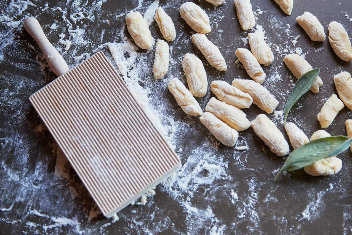 Ridged pasta board with rolling pin and fresh gnocchi dusted with flour on dark surface with sage leaves.