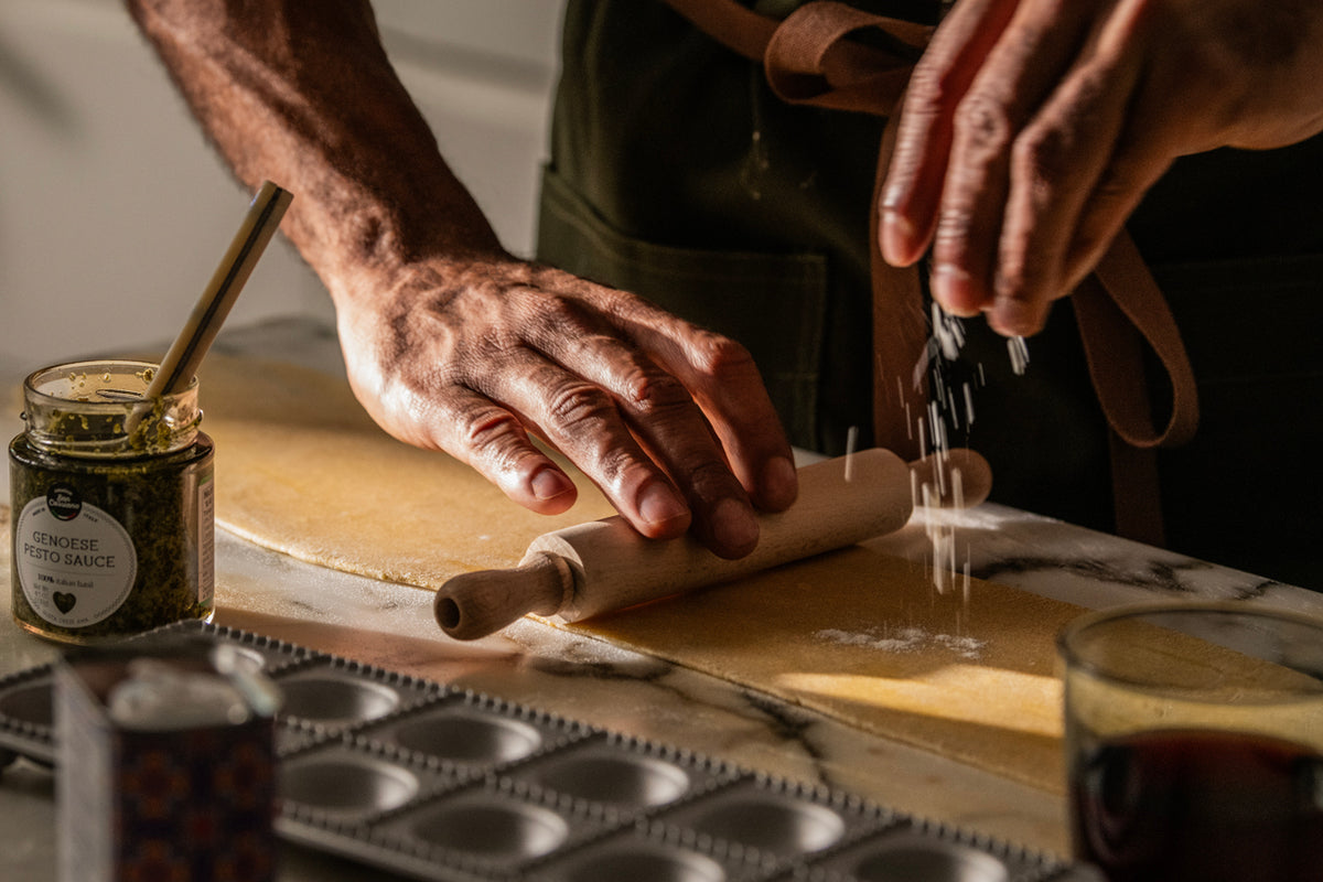 Hands using a wooden roller and metal ravioli mold to shape pasta dough on a work surface.