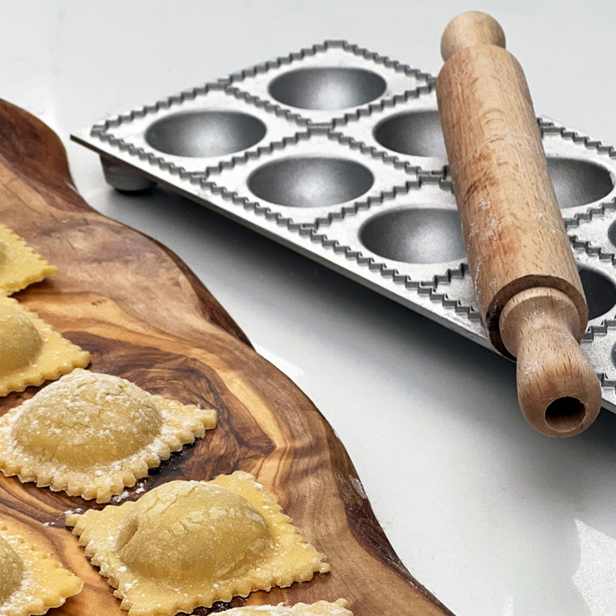 Metal ravioli mold with 12 cavities, wooden roller, and finished ravioli on wooden board.