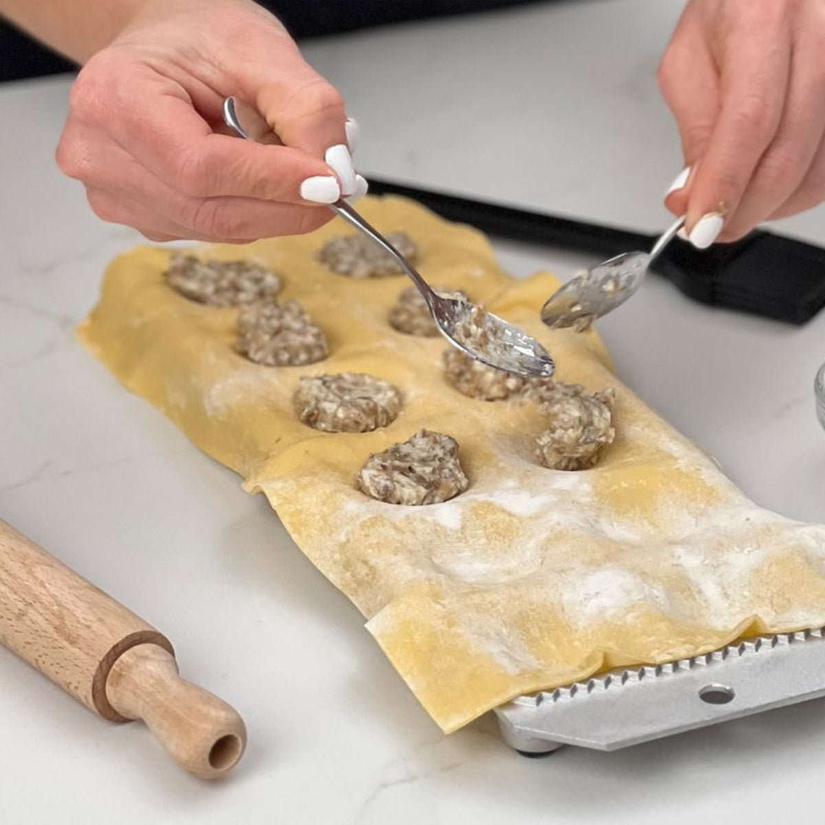 Hands filling pasta dough mold with meat filling using small spoon, with wooden roller and metal mold base visible.