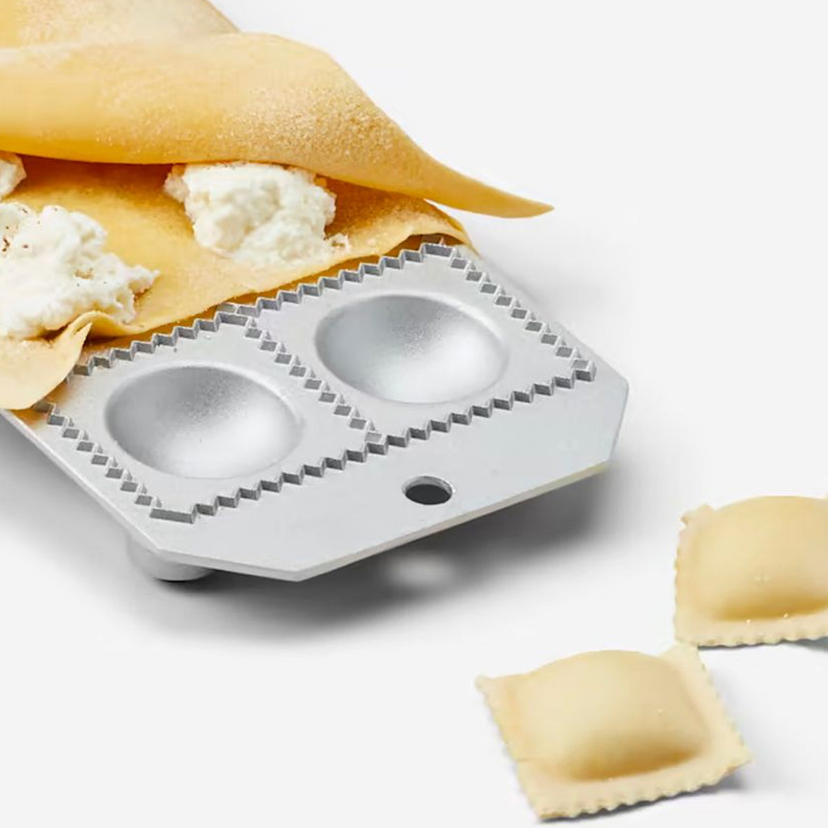 White plastic ravioli mold with two dome-shaped cavities, pasta sheet with filling, and finished ravioli pieces.