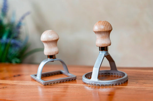 Two metal ravioli stamps with wooden handles displayed on a wooden surface.