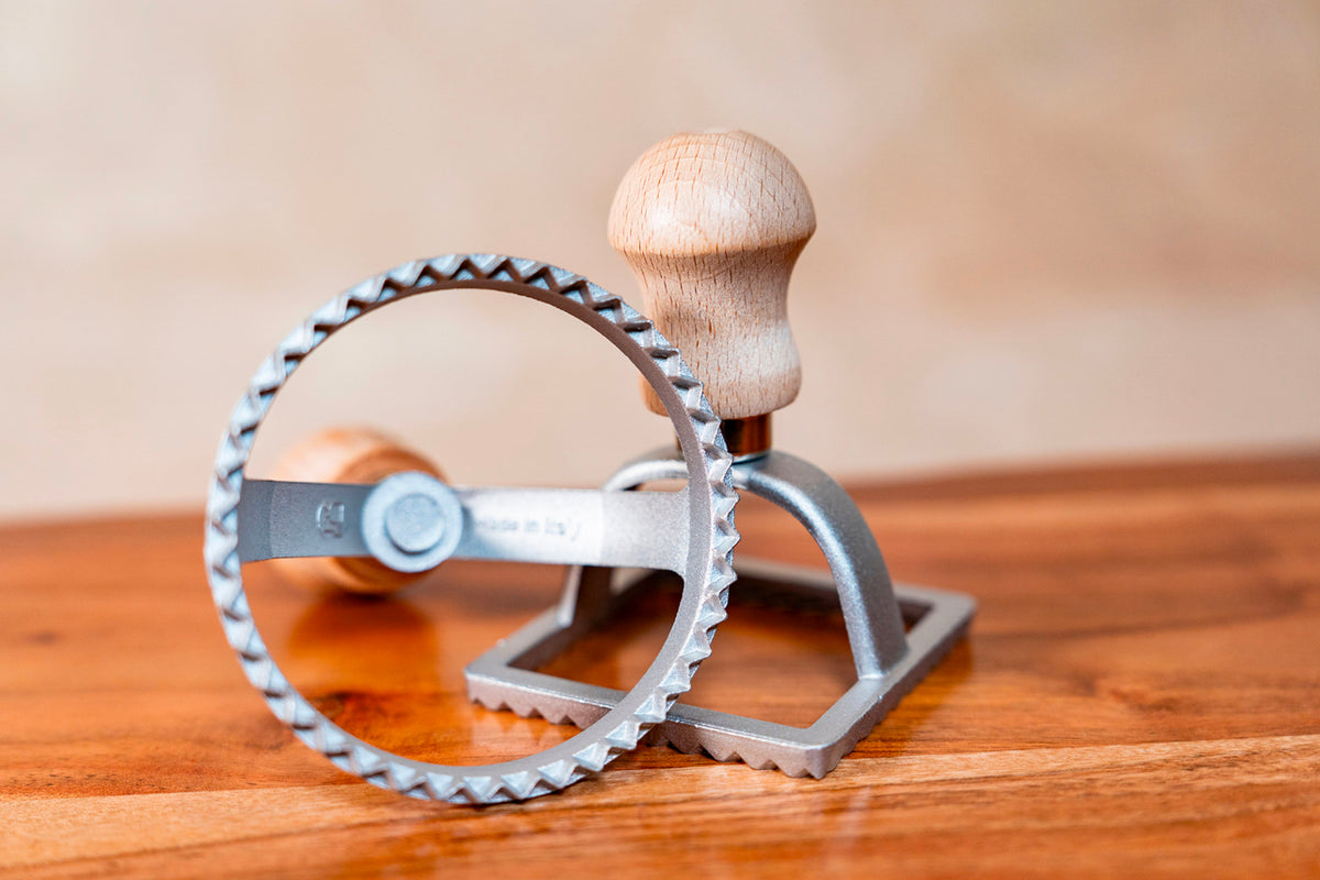 Metal ravioli stamp with scalloped circular cutting wheel and wooden handle on wooden surface.