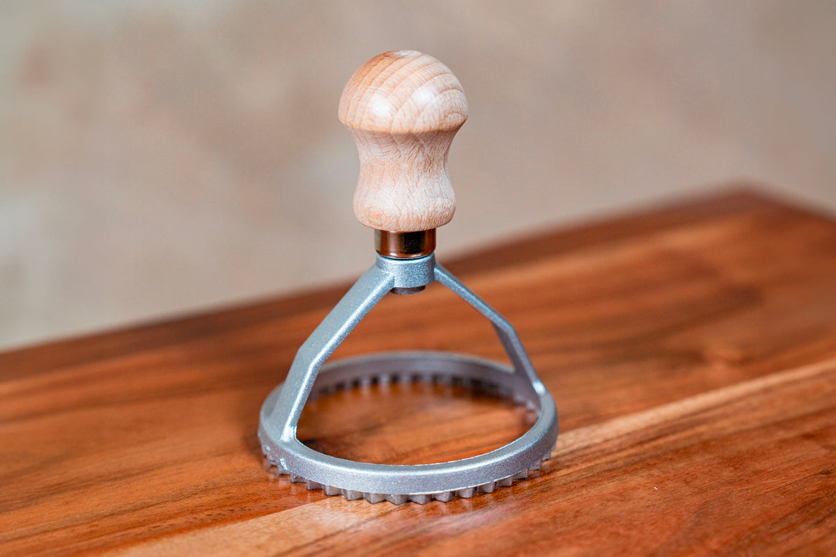 Wooden-handled ravioli stamp with metal circular cutting head and textured base, on wooden surface.