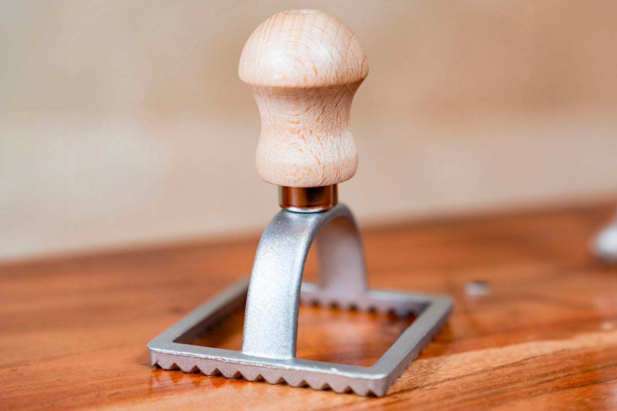 Wooden-handled metal ravioli stamp with square base and crimped edges on wooden surface.