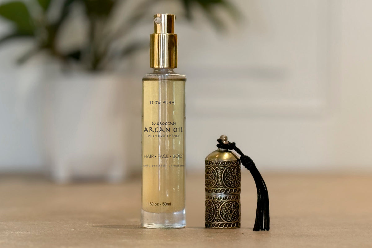 Clear glass bottle with gold spray top containing pale golden argan oil, labeled "100% Pure Moroccan Argan Oil," next to deco