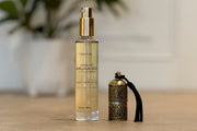 Clear glass bottle with gold spray top containing pale golden argan oil, labeled "100% Pure Moroccan Argan Oil," next to deco