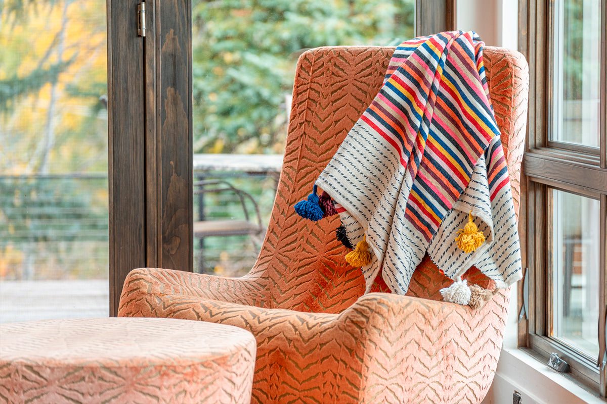 Striped Moroccan blanket with colorful tassels draped over an orange quilted chair by a window.