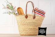 Woven straw tote basket with brown leather handles containing dried flowers, wooden spoon, and striped fabric.