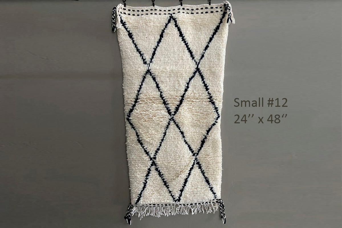 Cream wool rug with black diamond pattern and fringe edges, displayed flat against gray wall.
