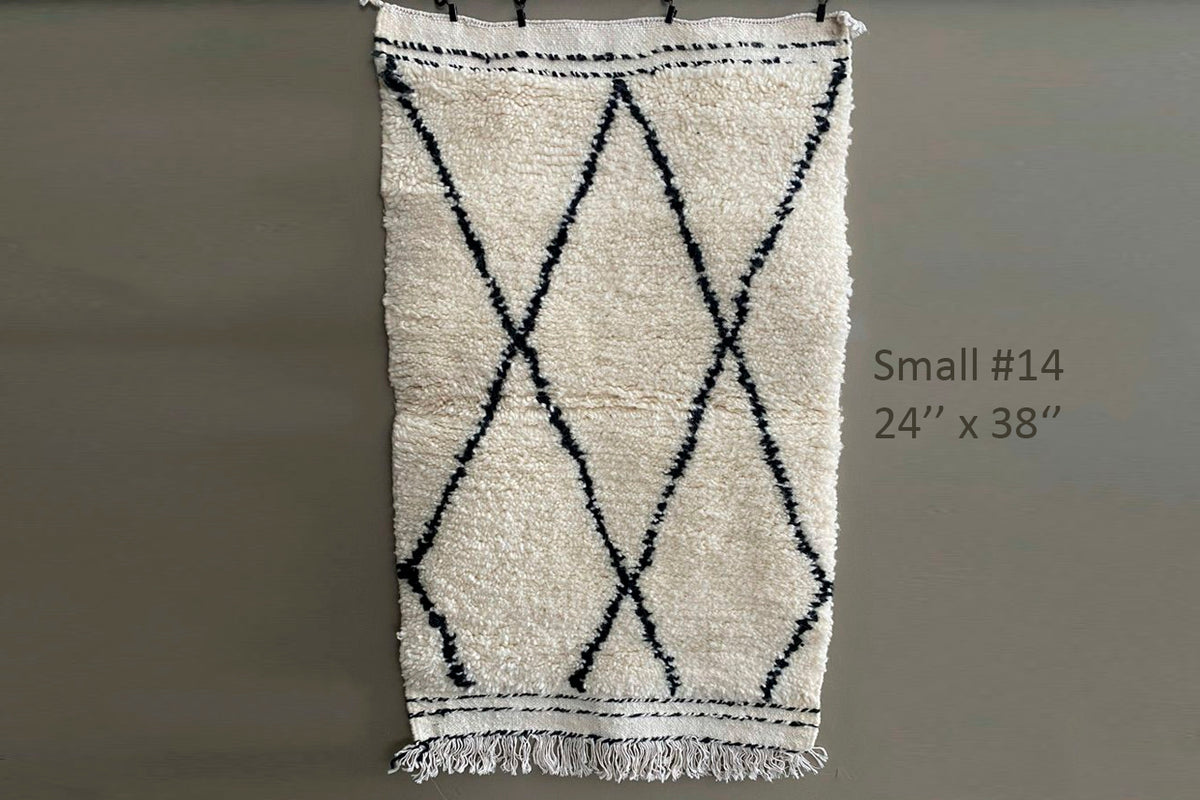 Cream wool rug with black diamond pattern and fringe ends, hanging against gray wall.
