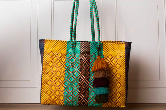 Woven tote bag with yellow, turquoise, and brown geometric pattern, green handles, and tassel embellishment.