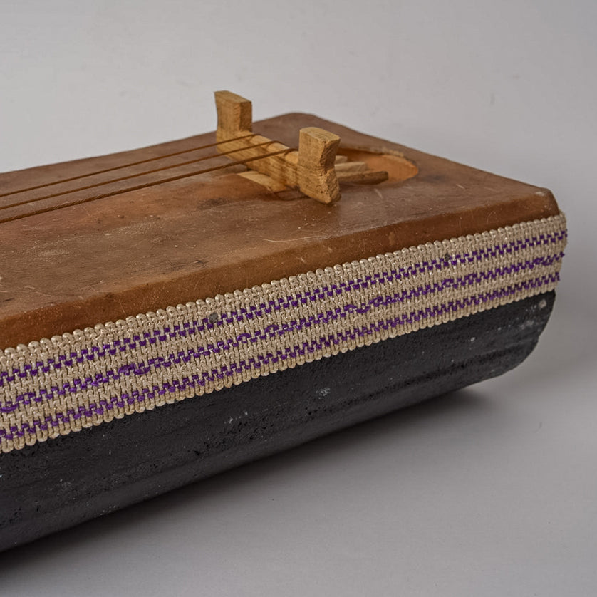 Wooden gimbri with three strings, wooden bridge, and woven purple, cream, and black fabric trim along the body.