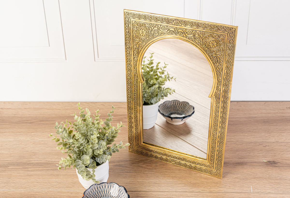 Gold rectangular mirror with ornate carved border and arched center opening, displayed on wooden surface next to potted plant