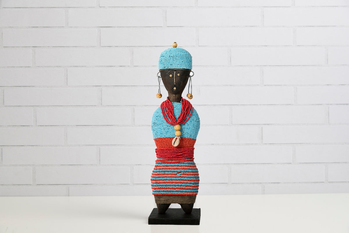 Handcrafted doll with turquoise knit hat, beaded earrings, red and striped fabric wrapping, and dark wooden base.