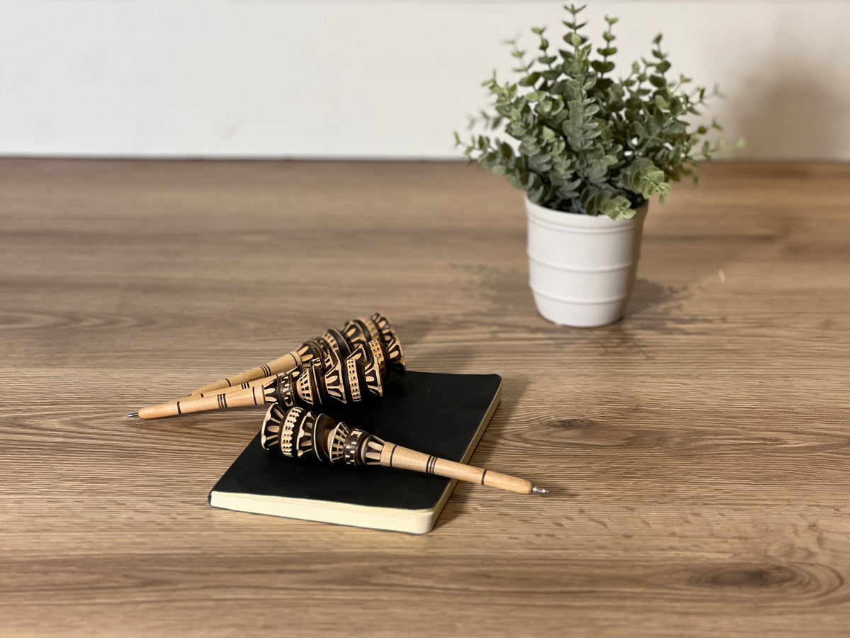 Two wooden pens with geometric black patterns on a black notebook beside a potted plant.
