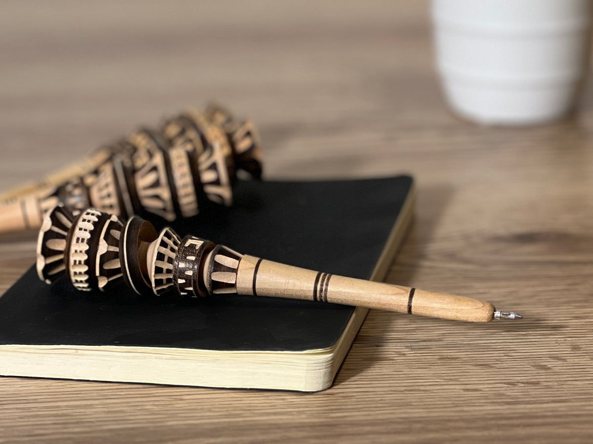 Wooden pen with carved geometric patterns on barrel, metal tip, resting on black notebook.