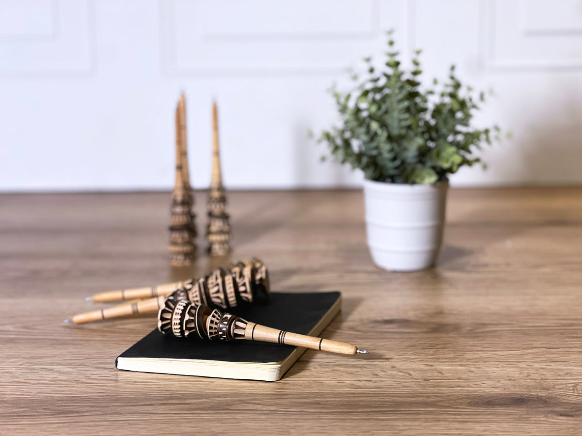 Pen with carved wooden barrel and decorative black patterned band resting on black notebook beside potted plant.