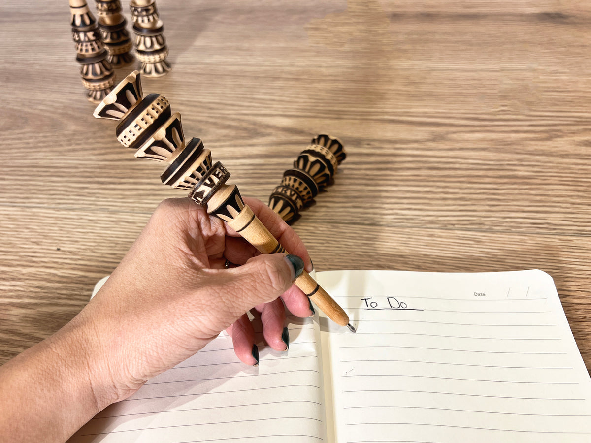 Hand holding decorative wooden pen with carved geometric patterns over open notebook on wooden surface.