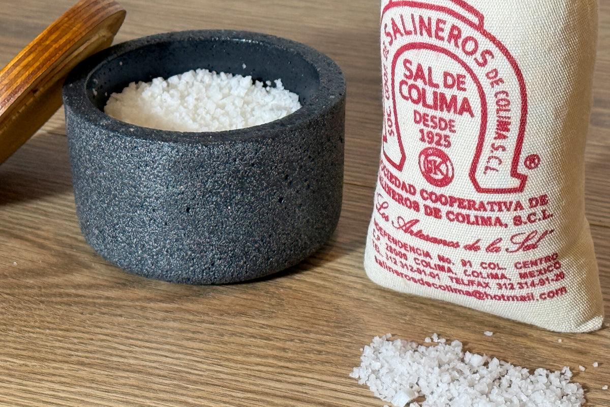 Coarse white sea salt in stone mortar with wooden pestle, cloth bag with red label, and scattered salt crystals on wooden sur