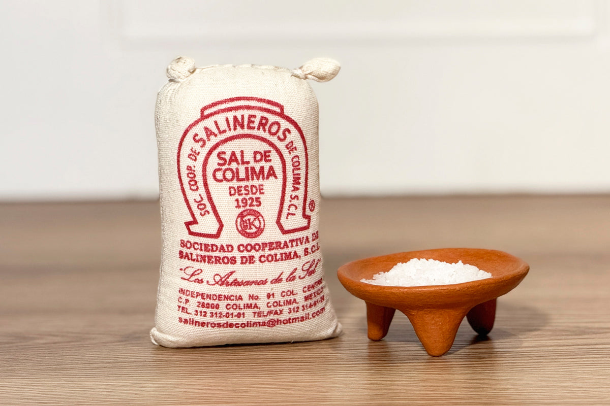 Cream-colored cloth bag with red "Sal de Colima" label next to wooden bowl containing white sea salt granules.