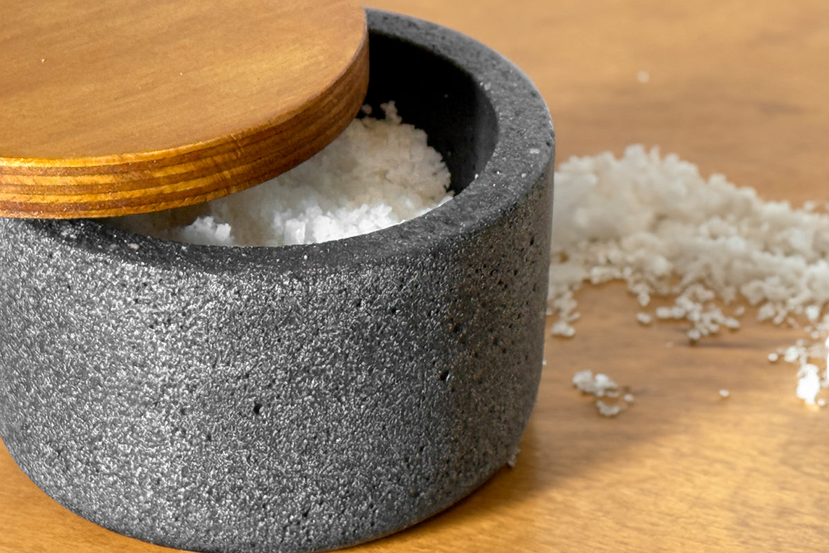 Gray concrete salt cellar with wooden lid, partially filled with white salt, on wooden surface.
