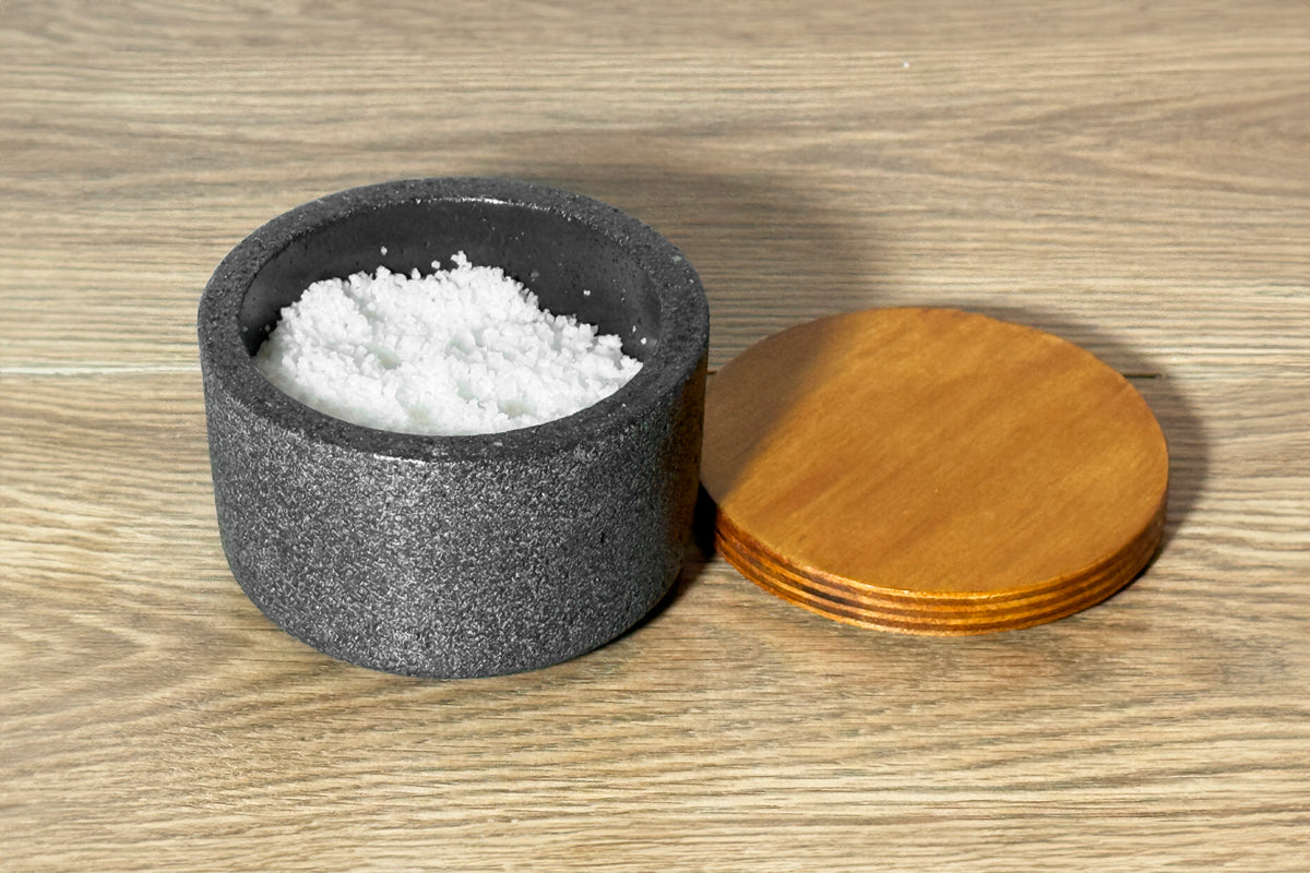 Dark gray concrete container filled with white salt and matching wooden lid on wooden surface.