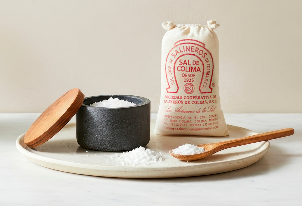 Black concrete salt cellar with wooden lid on ceramic tray, accompanied by wooden spoon and salt bag.