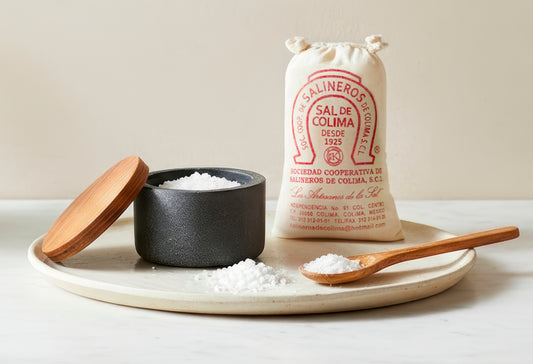 Black ceramic salt cellar with wooden lid and spoon, cream linen salt bag, and wooden serving spoon on ceramic plate.