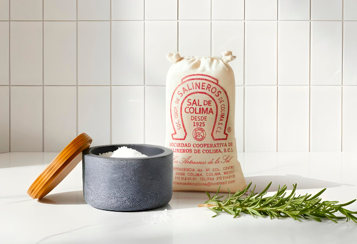 Gray stone salt cellar with wooden lid beside cream canvas bag of Colima sea salt and fresh rosemary sprig.