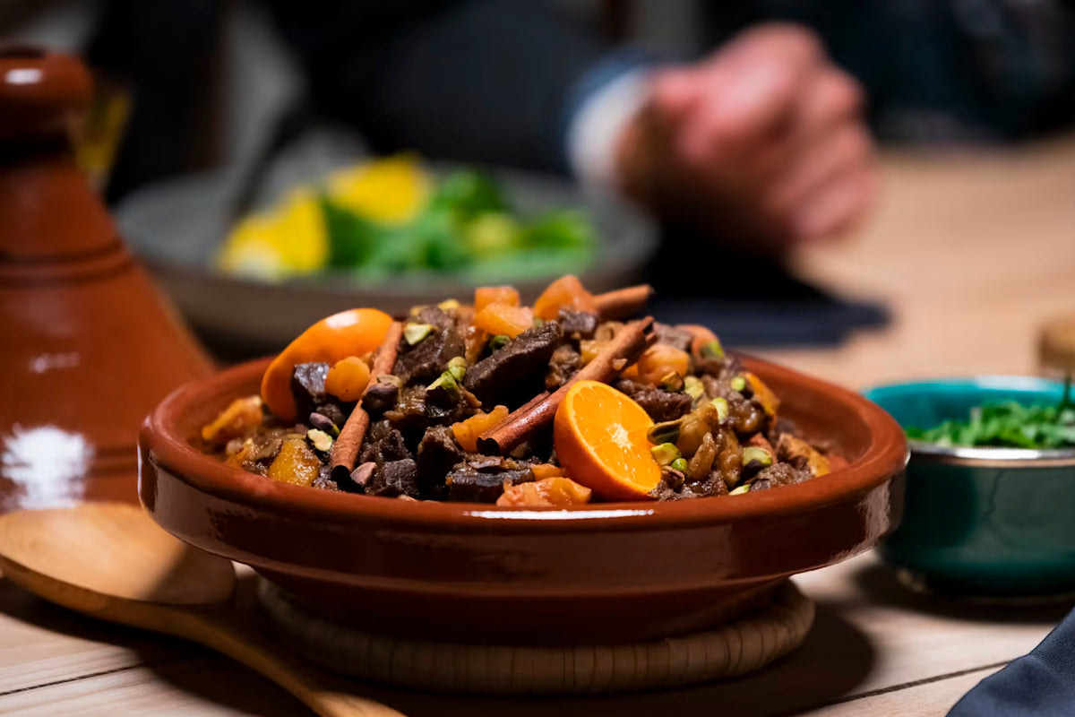 Terracotta tagine with cone-shaped lid containing braised meat and apricots, surrounded by serving bowls on wooden surface.