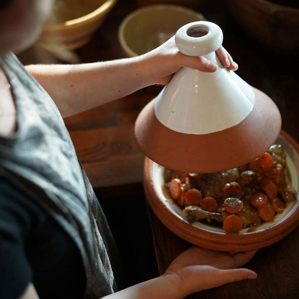 Terracotta tagine with white cone-shaped lid being held, containing cooked vegetables and meat inside.