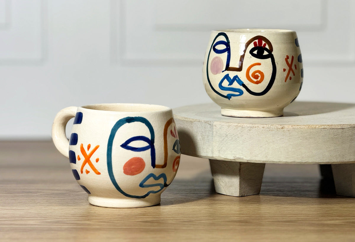 Two cream ceramic mugs with hand-painted abstract faces in blue, red, orange, and black lines.