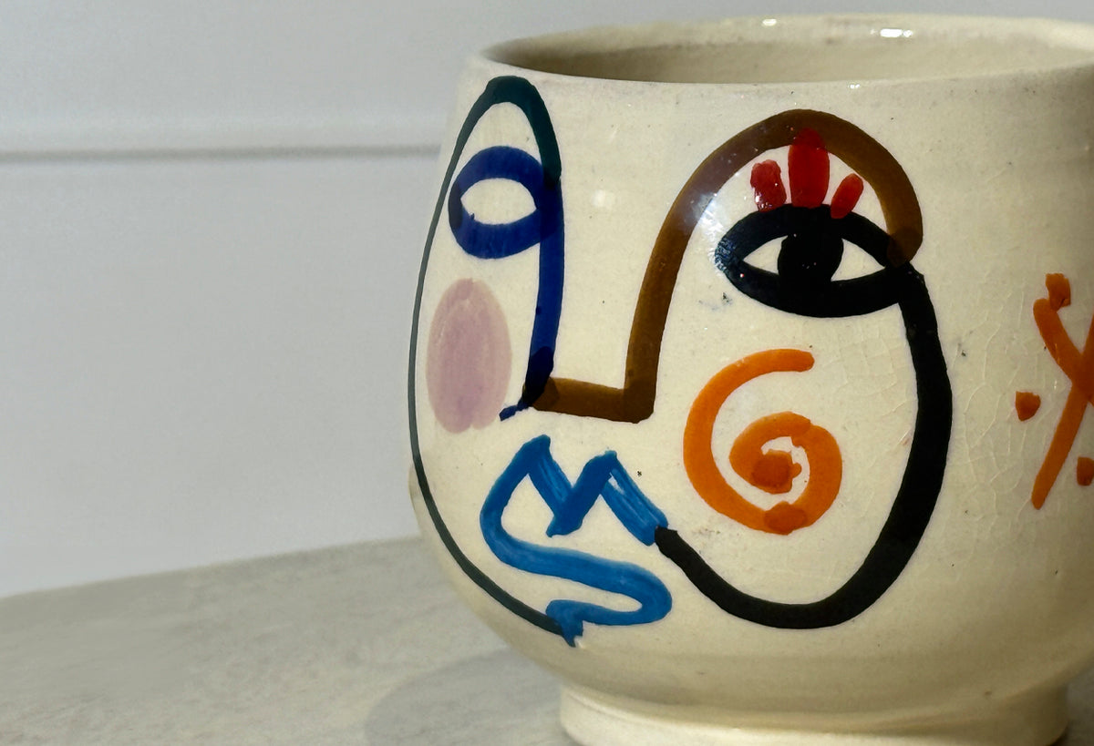 Ceramic mug with handpainted abstract face featuring blue, black, orange, and red line work on cream background.