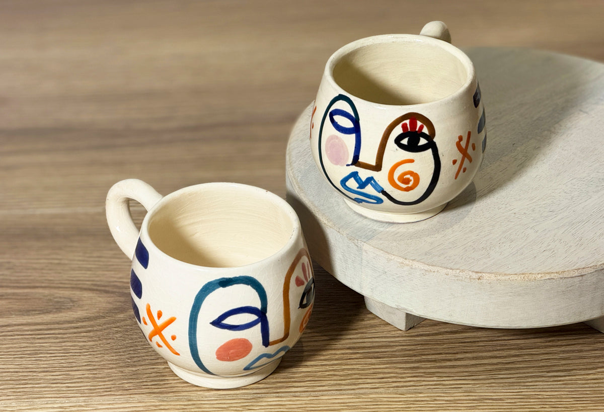 Two cream ceramic mugs with hand-painted abstract faces in blue, red, and orange lines on wooden surface.