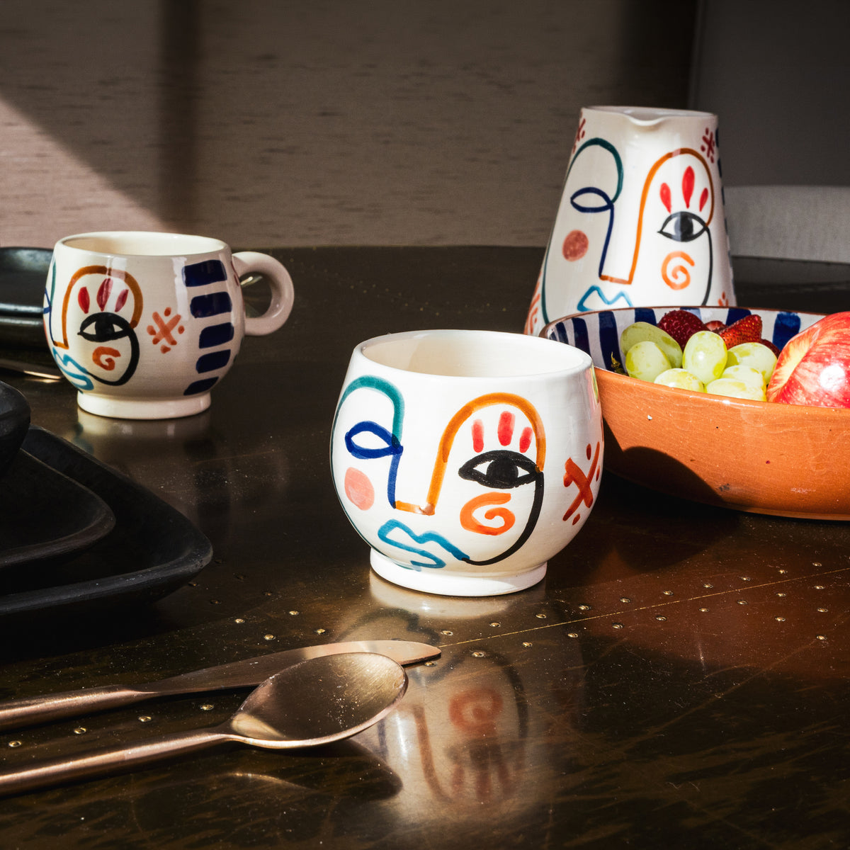Two ceramic mugs with hand-painted abstract faces in navy, orange, and red on cream background.
