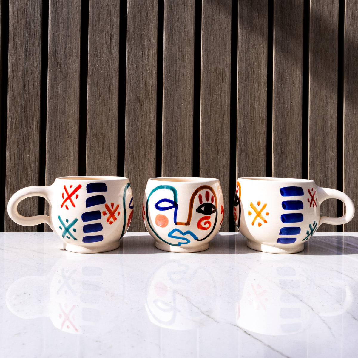 Three white ceramic mugs with hand-painted abstract faces and geometric patterns in blue, red, and yellow on marble surface.