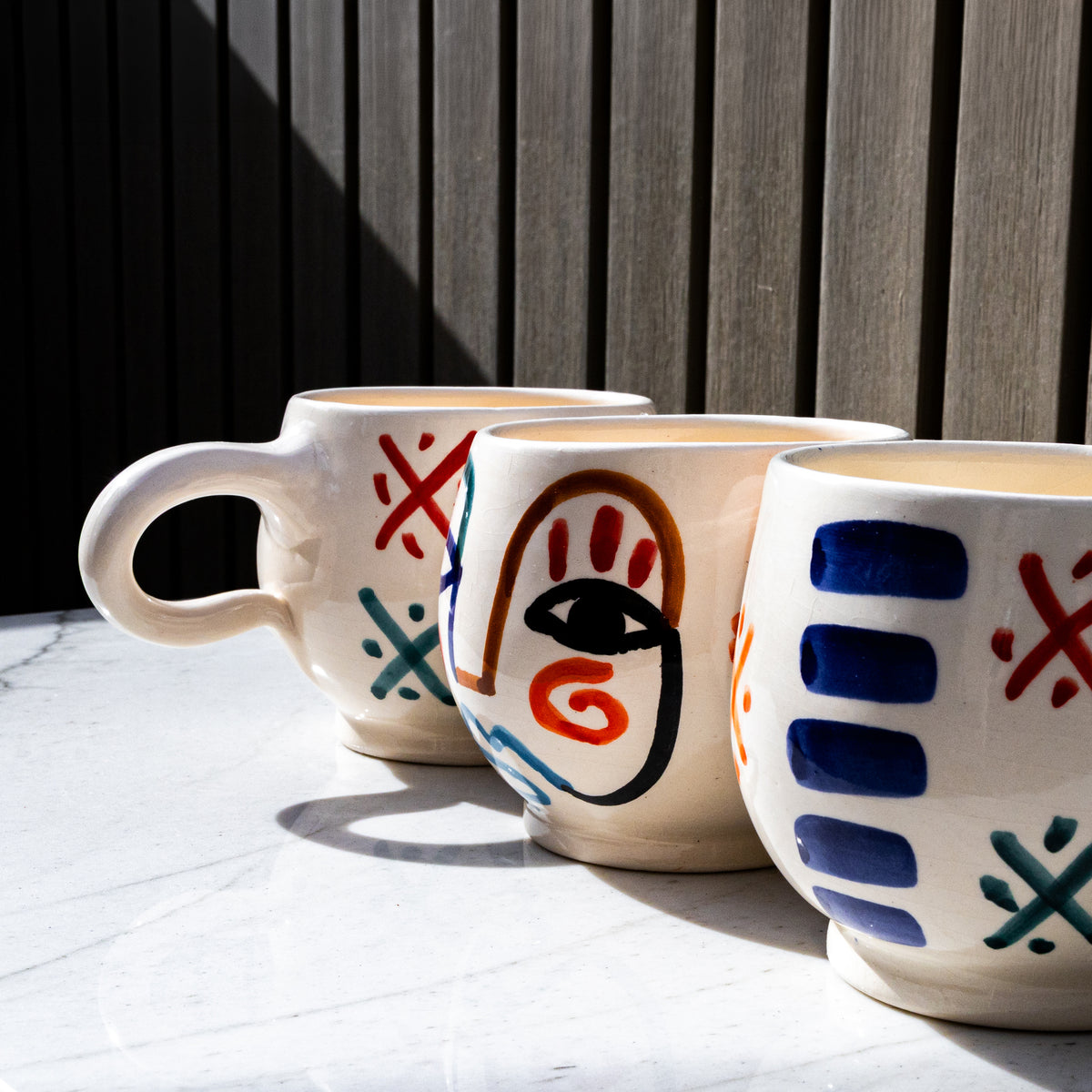 Two ceramic mugs with hand-painted abstract faces in red, blue, and teal designs on cream-colored bodies.