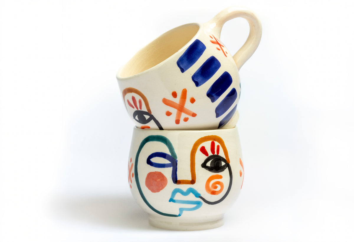 Two ceramic mugs with cream-colored bodies featuring hand-painted abstract faces in blue, orange, and black geometric designs